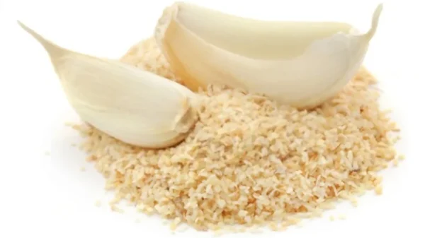 Close-up of two garlic cloves, one whole and one with its husk split open, resting on a mound of Dehydrated Garlic granules. The granules are evenly scattered, highlighting their texture and color contrast against the garlic cloves. The background is white.