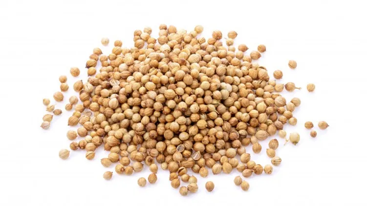 A pile of Coriander is scattered on a white background. The natural seeds are small, round, and light brown in color, with a slightly textured surface.