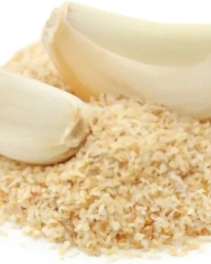 Close-up of two garlic cloves, one whole and one with its husk split open, resting on a mound of Dehydrated Garlic granules. The granules are evenly scattered, highlighting their texture and color contrast against the garlic cloves. The background is white.