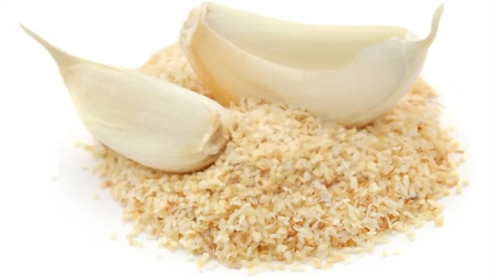 Close-up of two garlic cloves, one whole and one with its husk split open, resting on a mound of Dehydrated Garlic granules. The granules are evenly scattered, highlighting their texture and color contrast against the garlic cloves. The background is white.